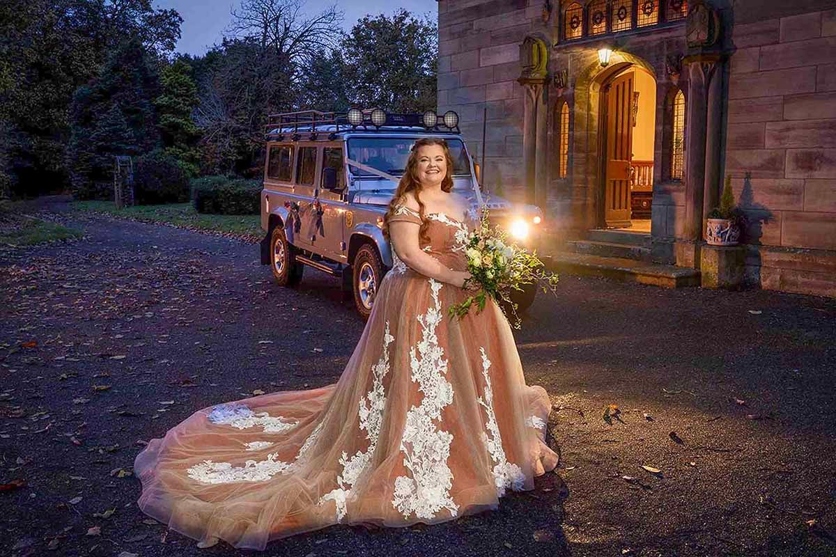Bride in champagne floral ball gown holding bouquet stands before a Land Rover at dusk outside historic Lady Gillford's House, Carlisle
