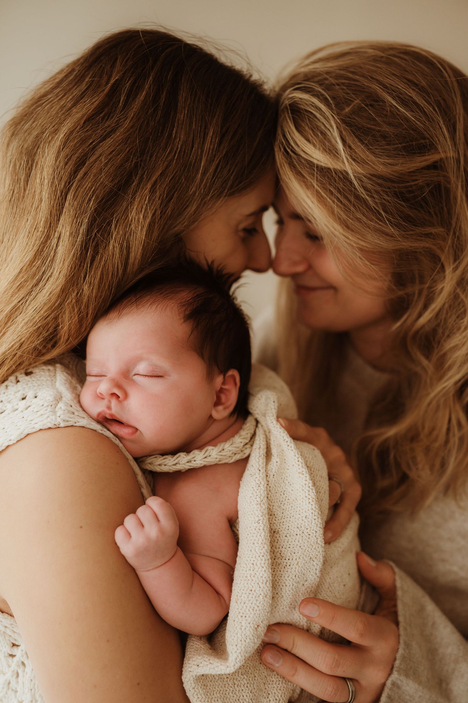 Two women tenderly touch foreheads while cradling a sleeping newborn baby wrapped in a cream knit blanket.