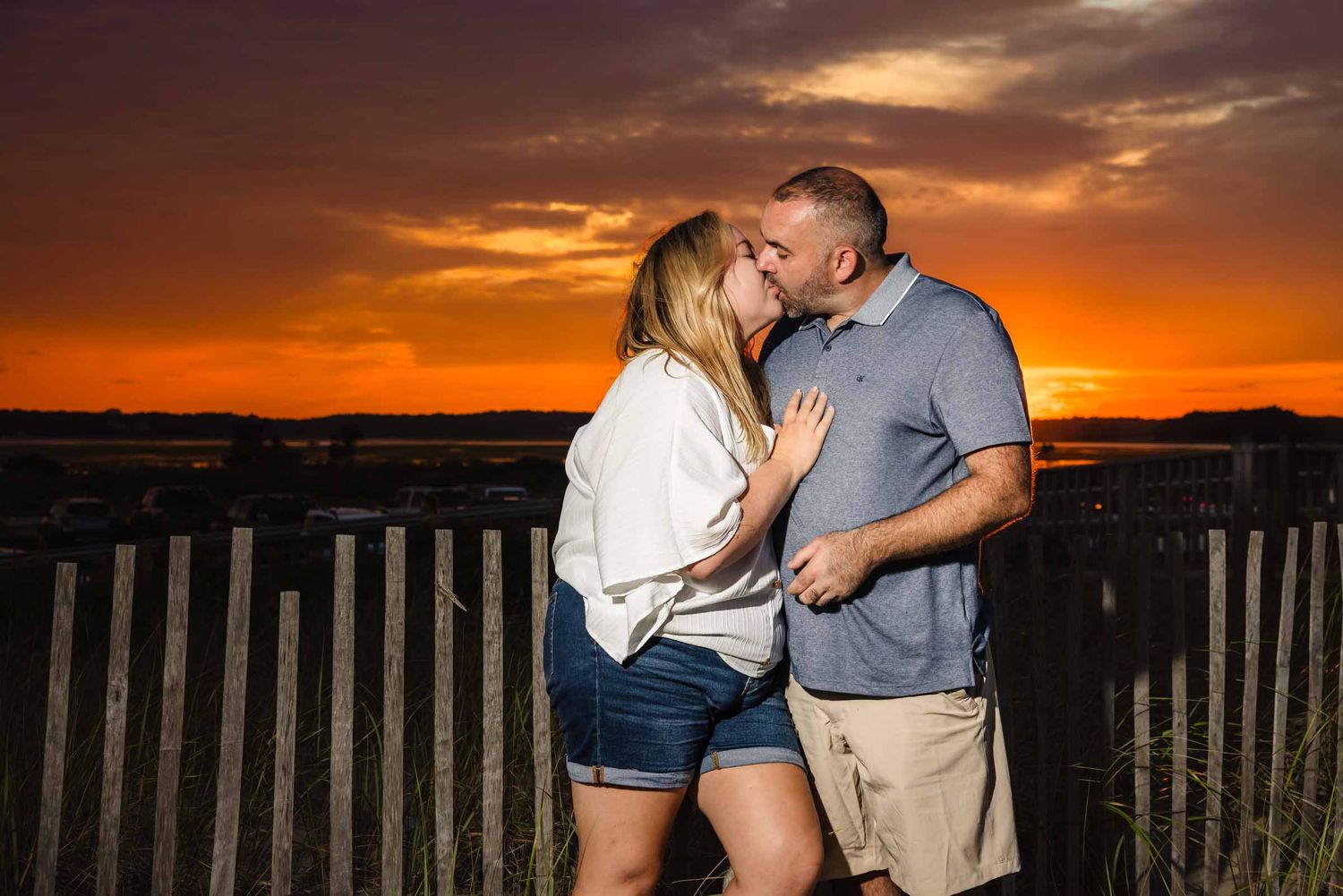 Couple sharing a romantic kiss in front of a wooden fence at sunset with a vivid orange sky backdrop.