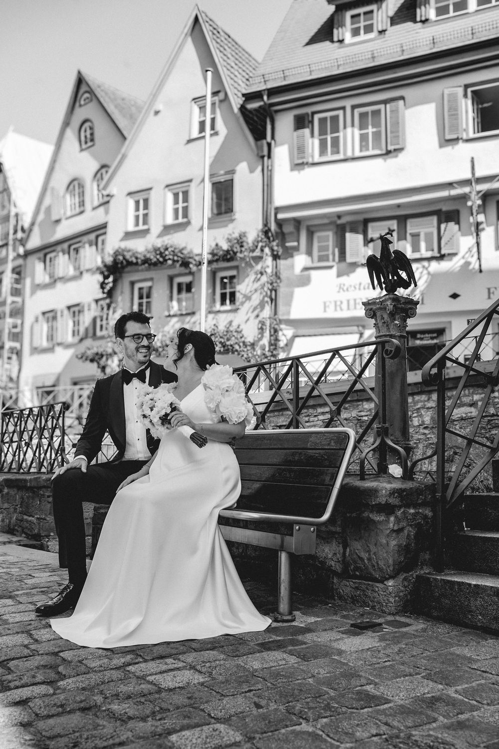 Black and white photo of a bride and groom sitting on a bench in a charming European town square on their wedding day.