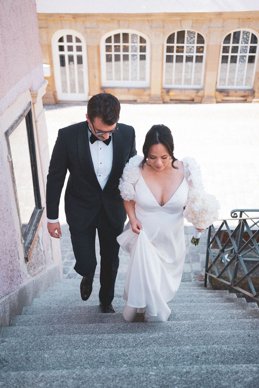 Bride and groom walking up stone steps, she in white gown with bouquet, he in black tuxedo with bow tie.