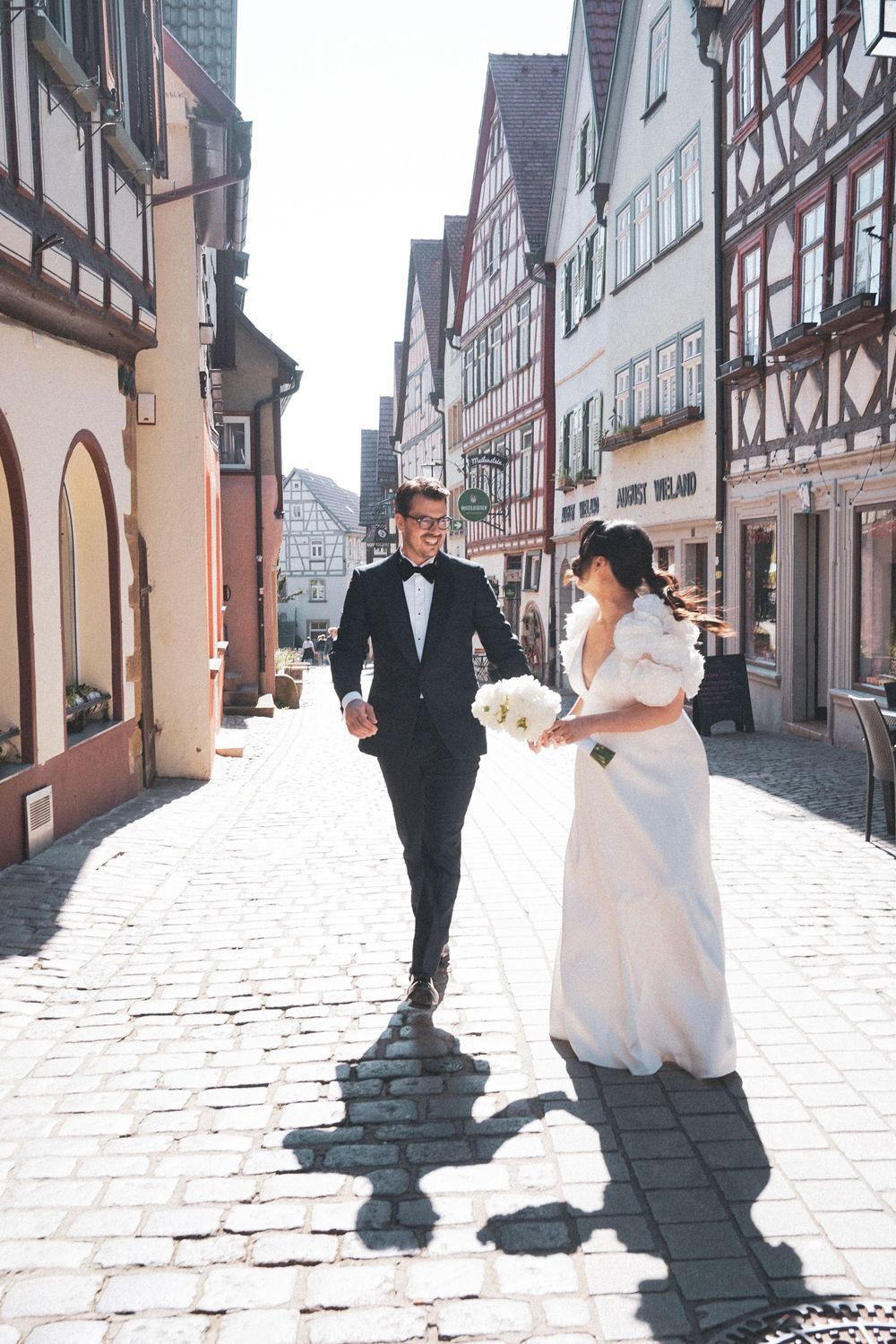 Bride and groom walk hand in hand on cobblestone street lined with traditional half-timbered buildings on their wedding day.