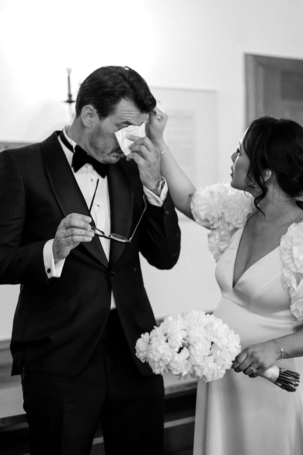 Black and white photo of groom crying while bride tenderly wipes his tears during emotional wedding ceremony.