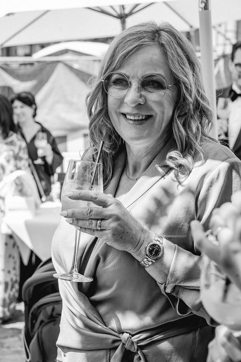 Smiling woman holding a wine glass at an outdoor event, wearing glasses and a watch, in black and white.