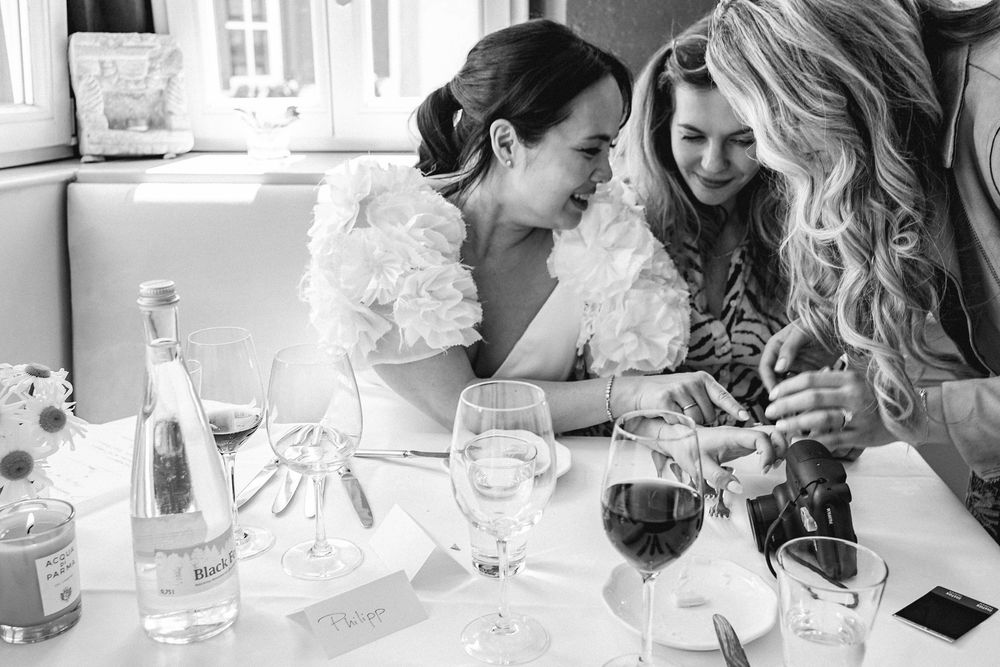 Two women in elegant attire share a joyful moment at a wedding reception table with wine glasses.