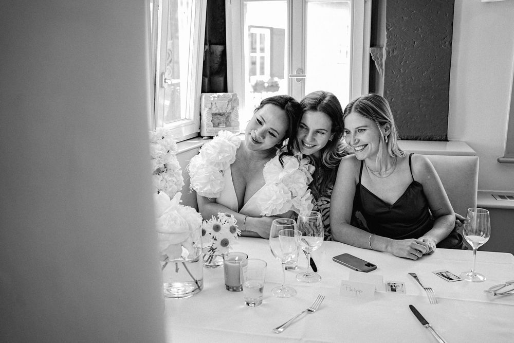 Three women laughing and embracing at a wedding reception table in black and white photography.