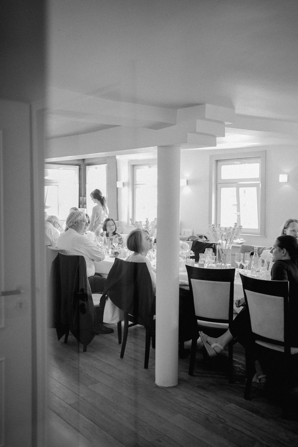Black and white photo of elegantly dressed guests dining in a bright restaurant with white columns and large windows.
