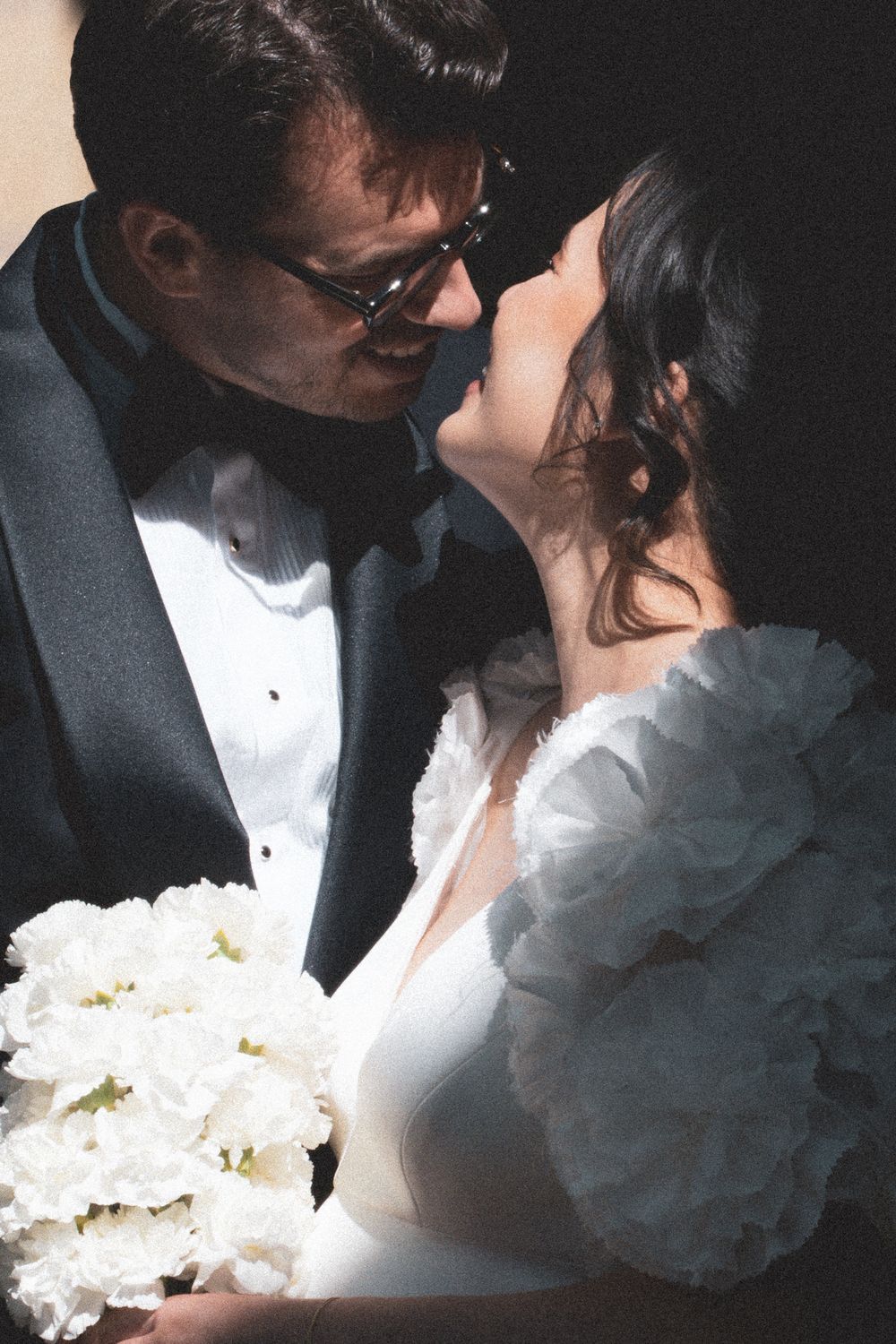 A bride and groom share an intimate moment, she holds white florals, he wears a tuxedo with bow tie.