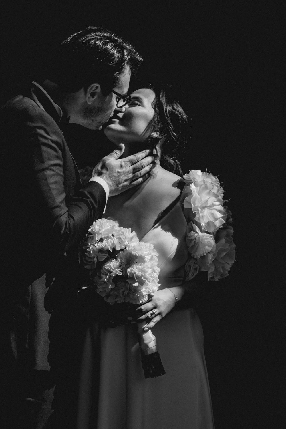 Black and white wedding photo of groom tenderly holding bride's face as they share a romantic kiss, bride holding bouquet.