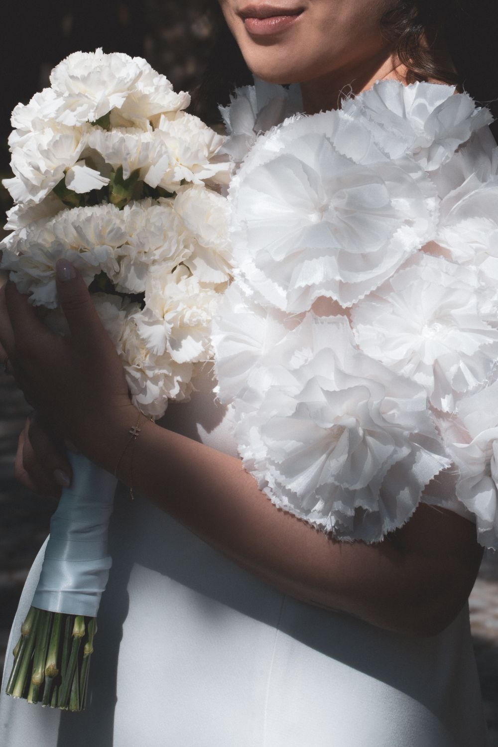 Bride in a ruffled white wedding dress holding a bouquet of white carnations in soft natural light.