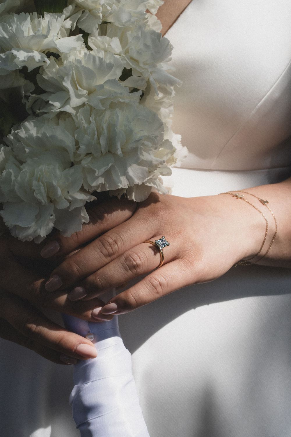 Bride holding white floral bouquet with elegant diamond engagement ring and gold bracelet in soft natural light.
