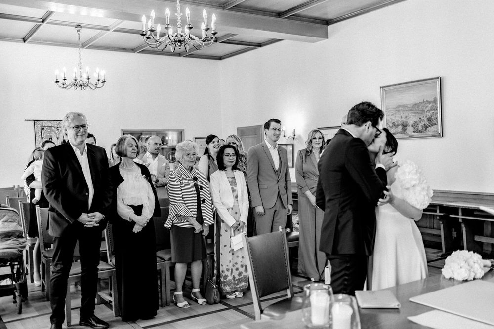 Black and white wedding ceremony photo showing couple kissing at altar with guests watching in elegant room.