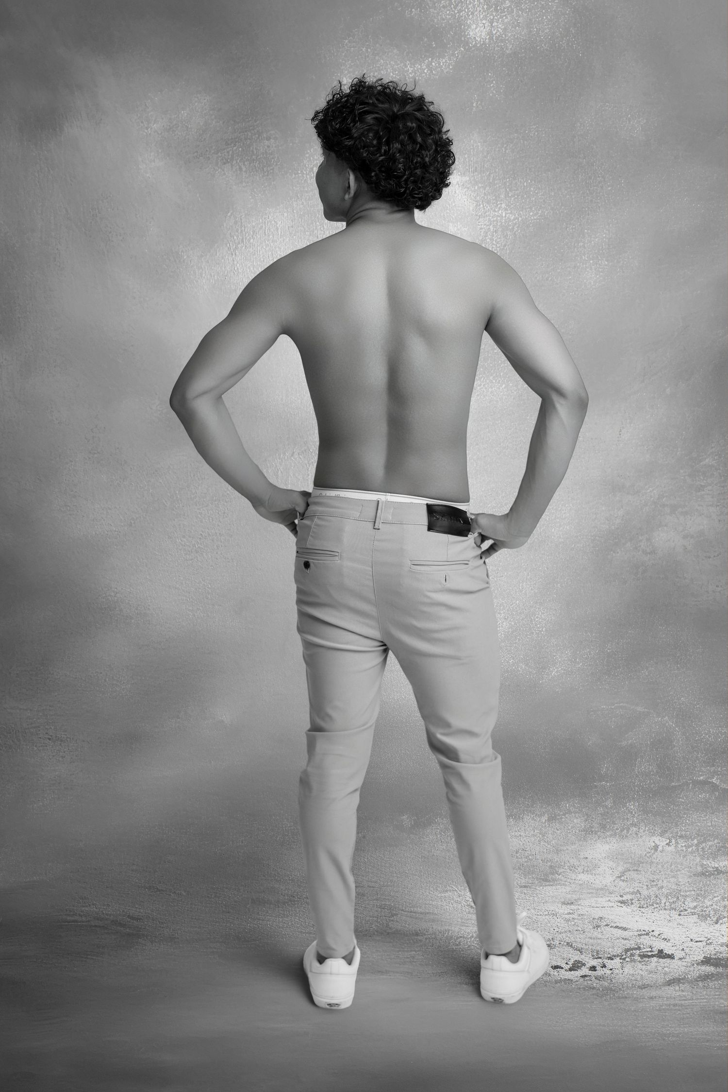 Black and white photo of a shirtless man with curly hair posing with hands on hips, wearing light trousers and white sneakers.