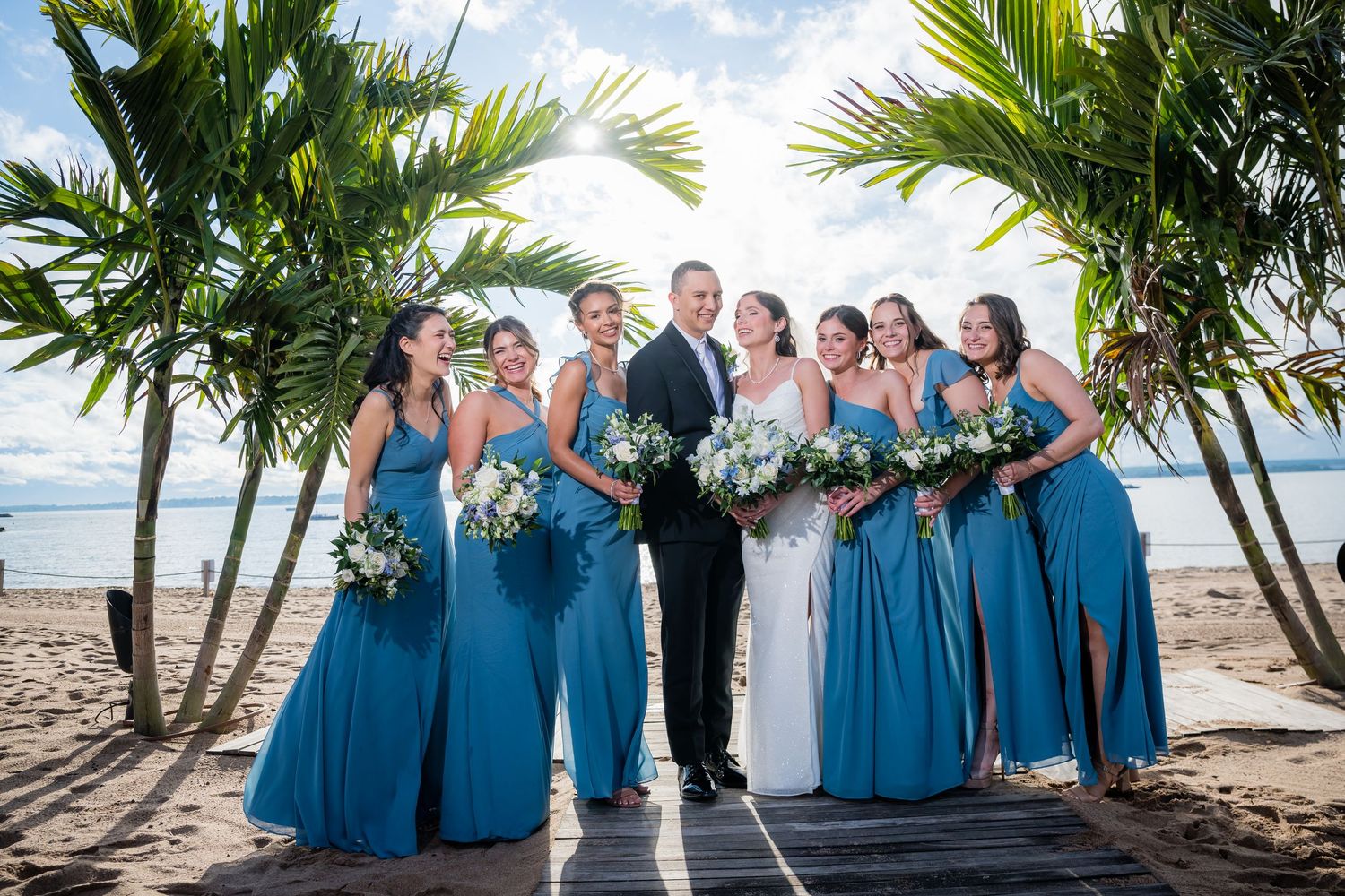 Bride and groom pose with bridesmaids in blue gowns holding white bouquets on a tropical beach wedding venue.