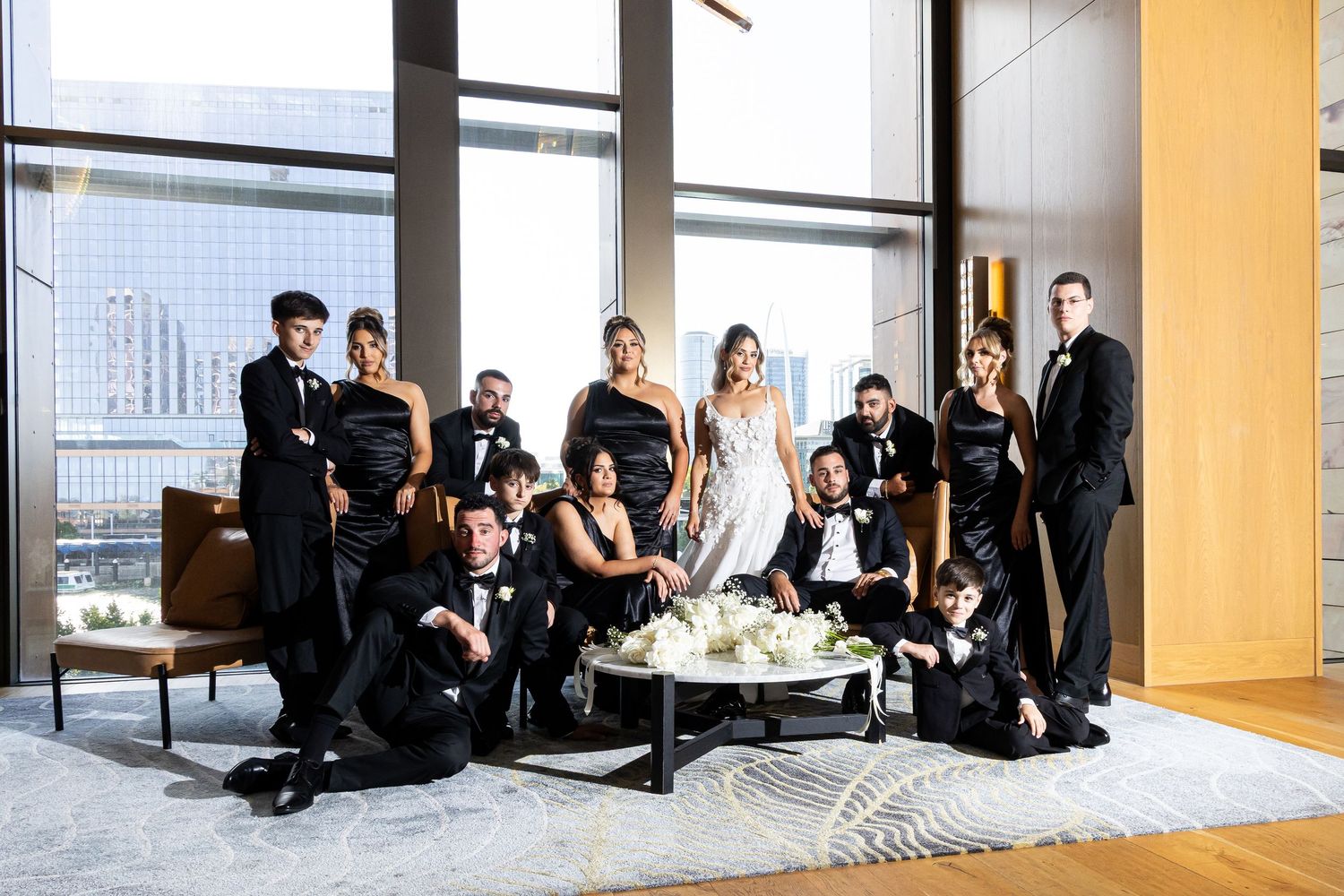Wedding party in black formal attire poses elegantly in a modern venue with floor-to-ceiling windows.