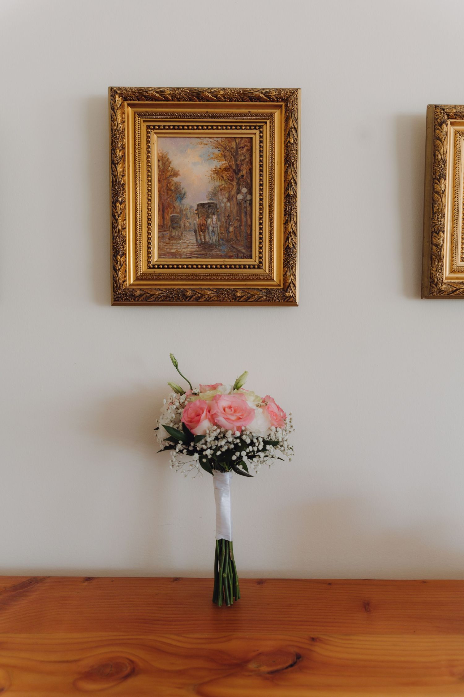Elegant bridal bouquet of pink roses and baby's breath on a wooden table below framed artwork.