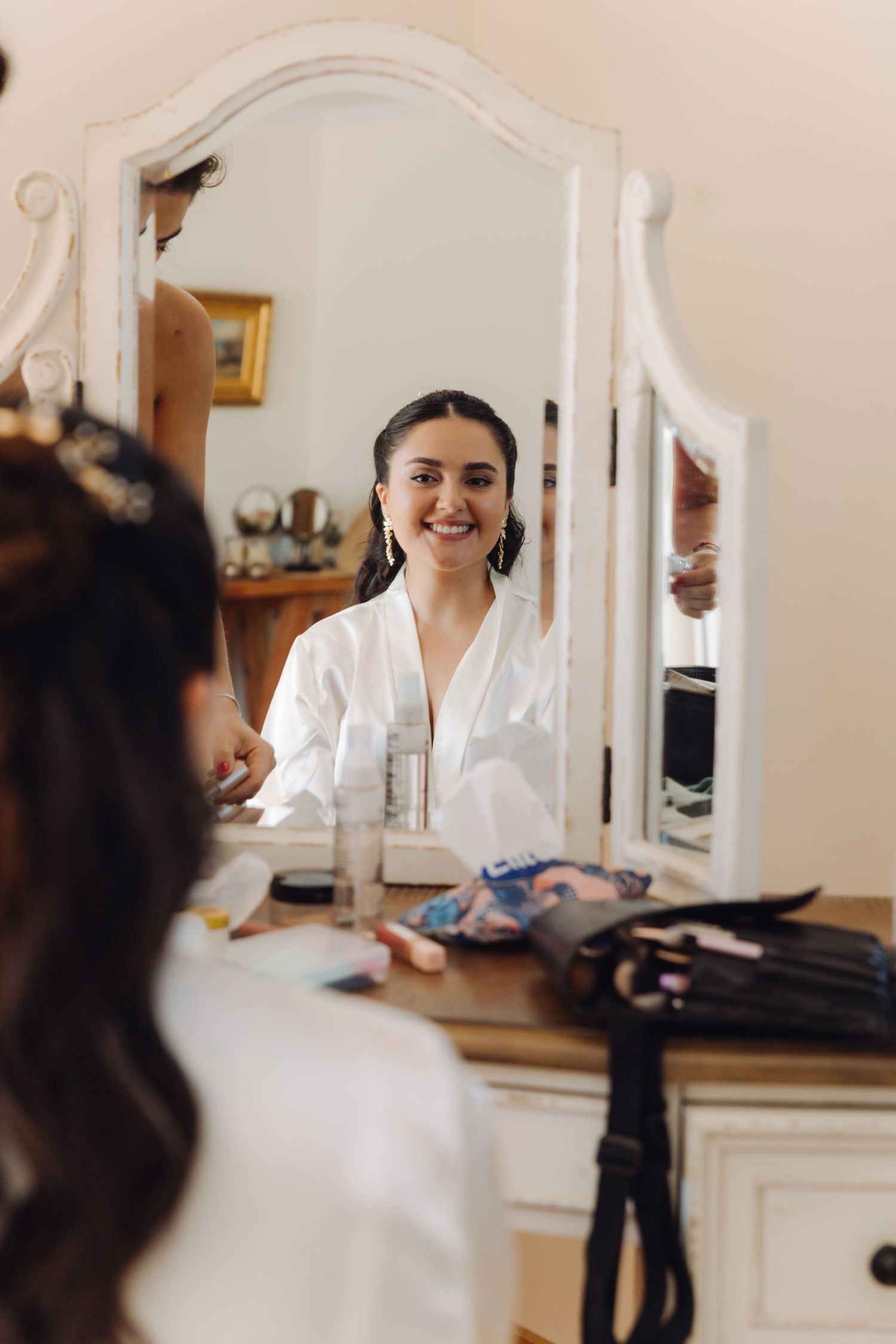 Beautiful bride in white robe smiling at her reflection in a vintage mirror during wedding prep.