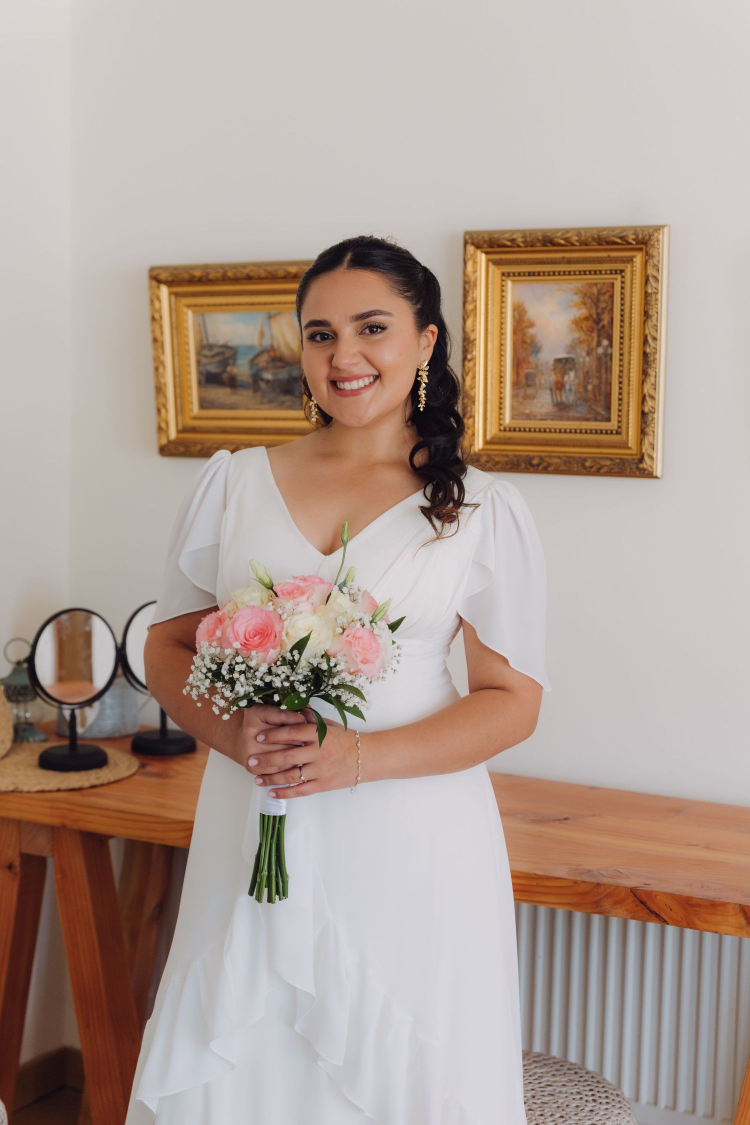 Radiant bride in white V-neck wedding dress holding pink rose bouquet in front of gold-framed paintings.