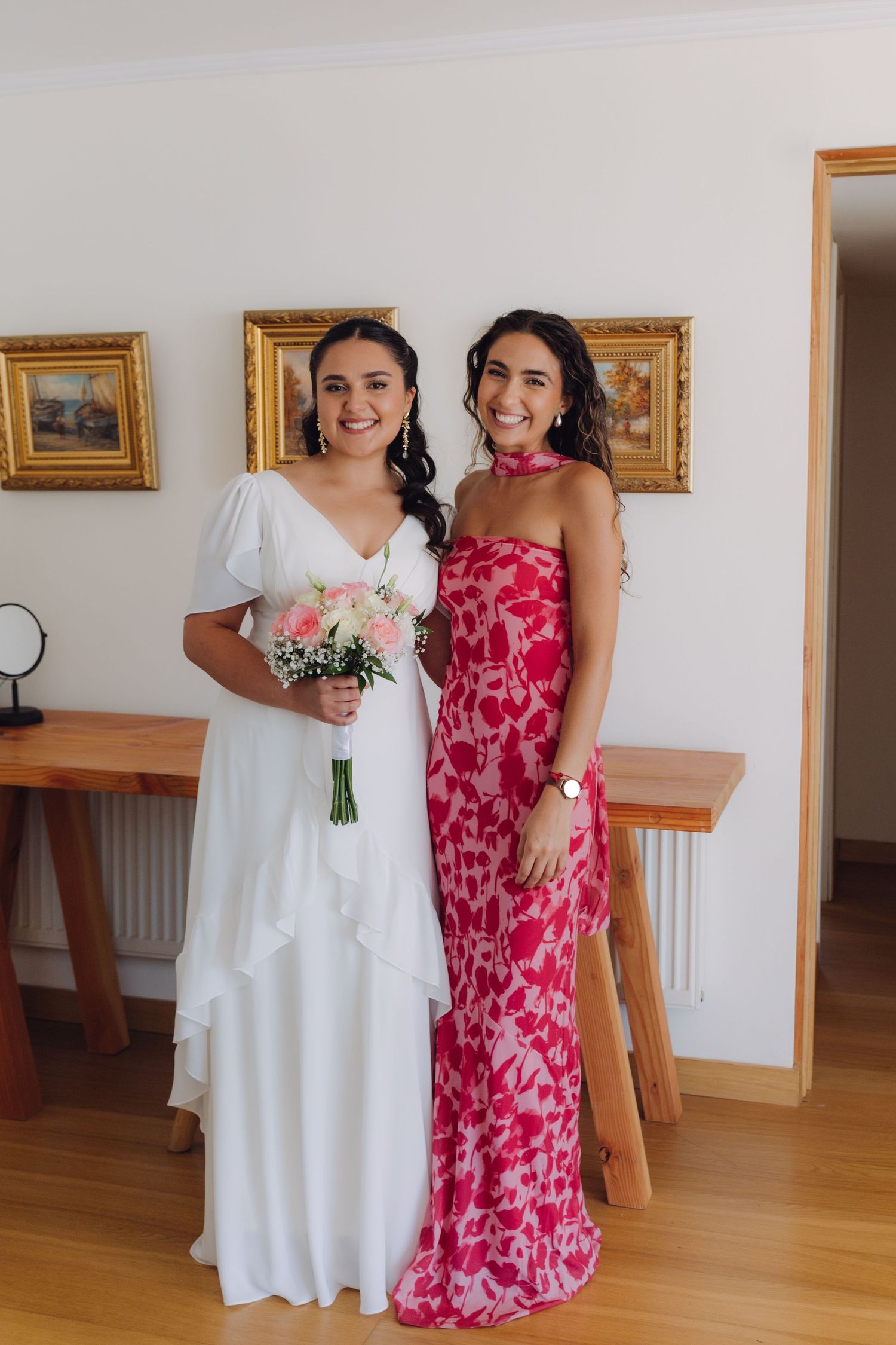 Smiling bride in white dress with pink rose bouquet posing with bridesmaid in pink floral gown.