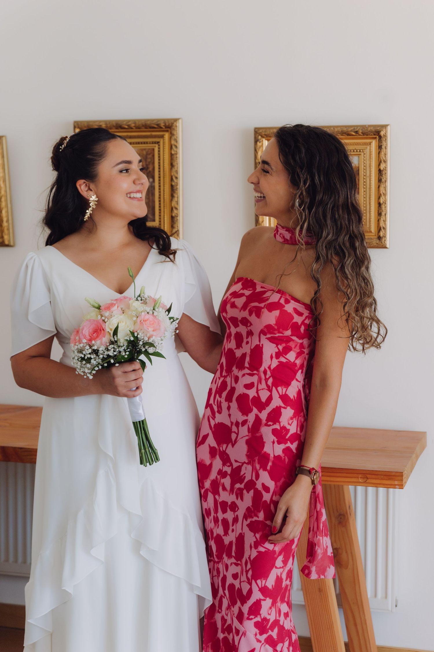 Bride in white dress holding pink bouquet smiles at bridesmaid in red floral dress indoors.