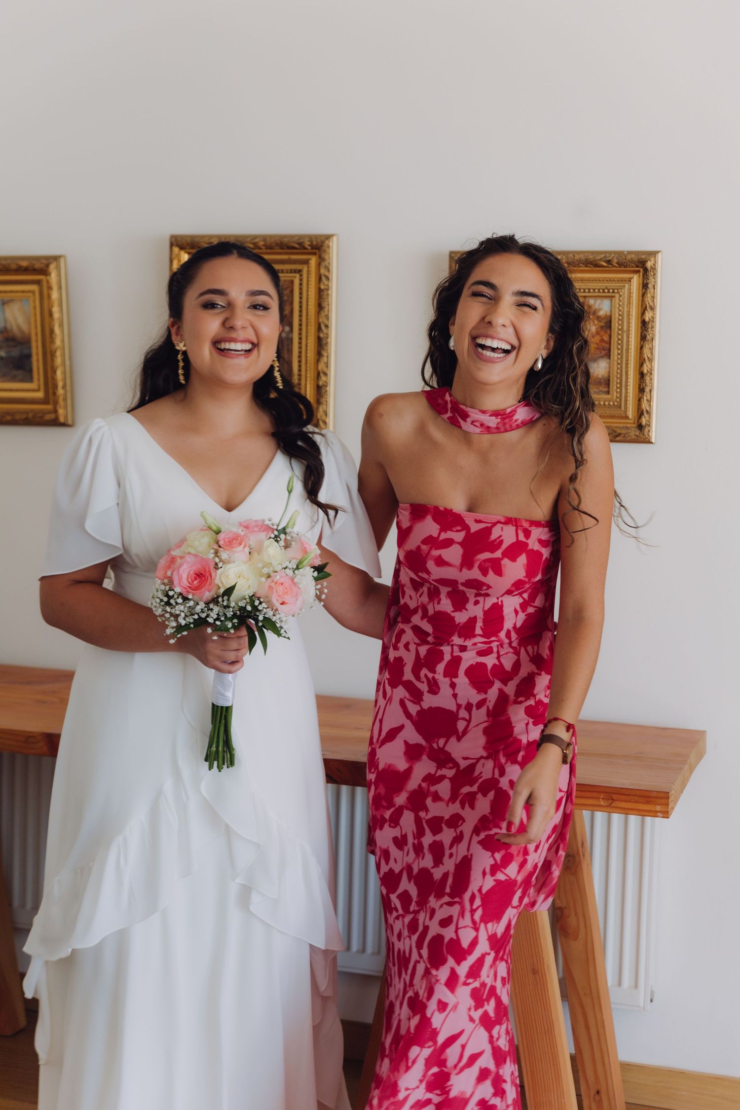 Bride and bridesmaid laughing joyfully together, bride holding pink bouquet in white wedding dress.