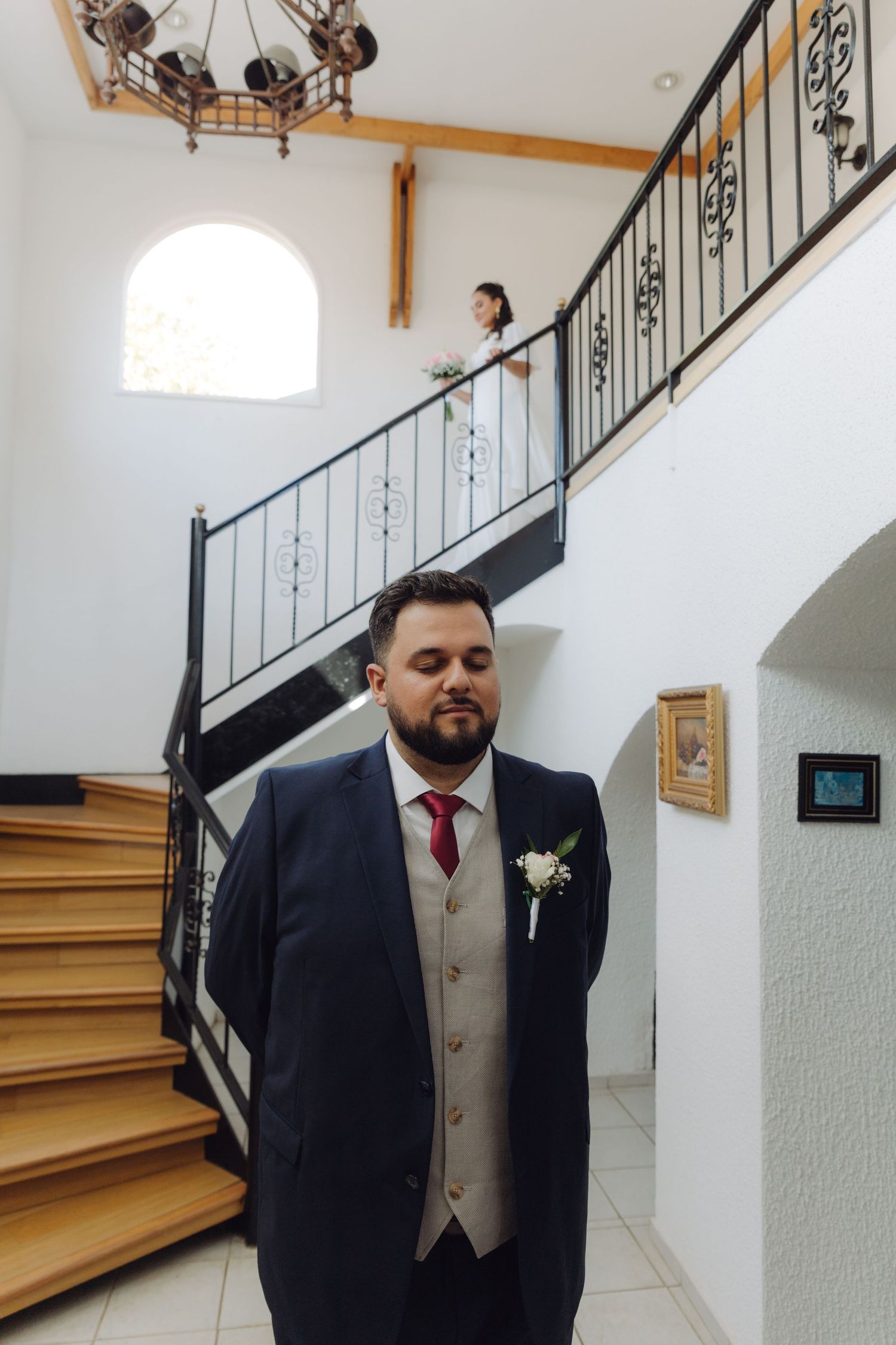 Groom in navy suit waits at staircase as bride descends in white dress for first look.