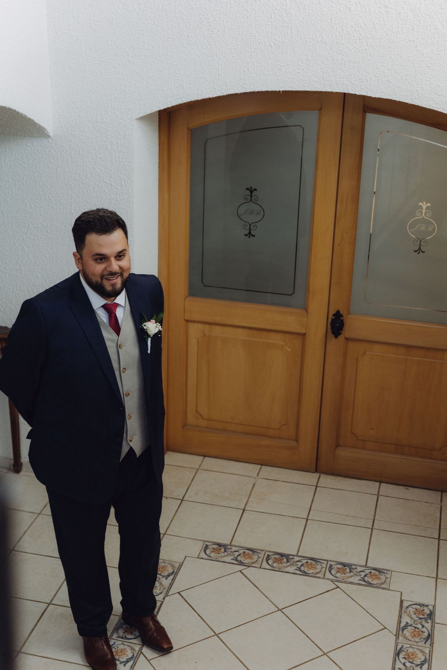 Groom in navy suit with boutonniere smiles standing by wooden doors at wedding venue.