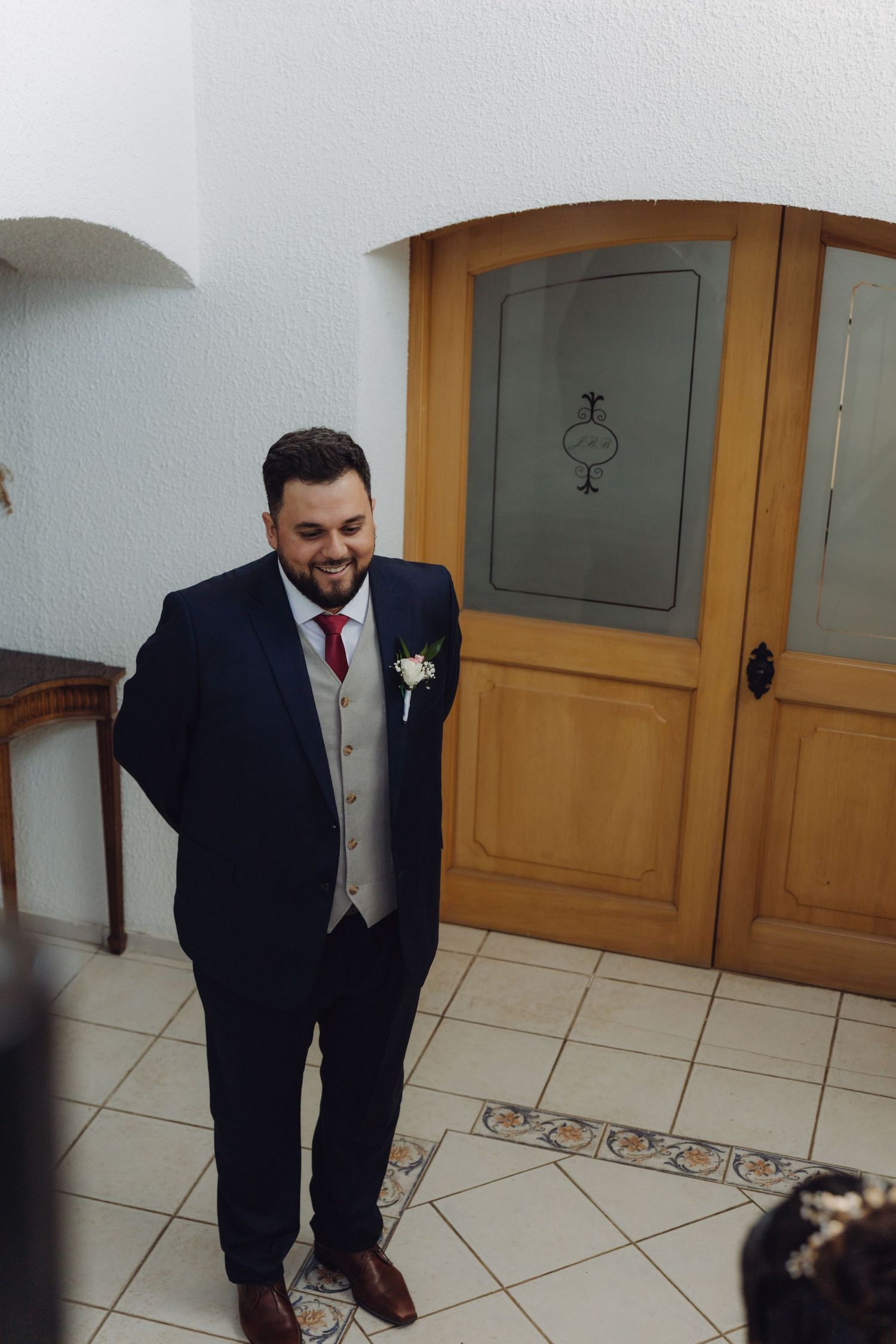 Groom in navy suit smiling downward near ornate wooden doors at wedding ceremony location.