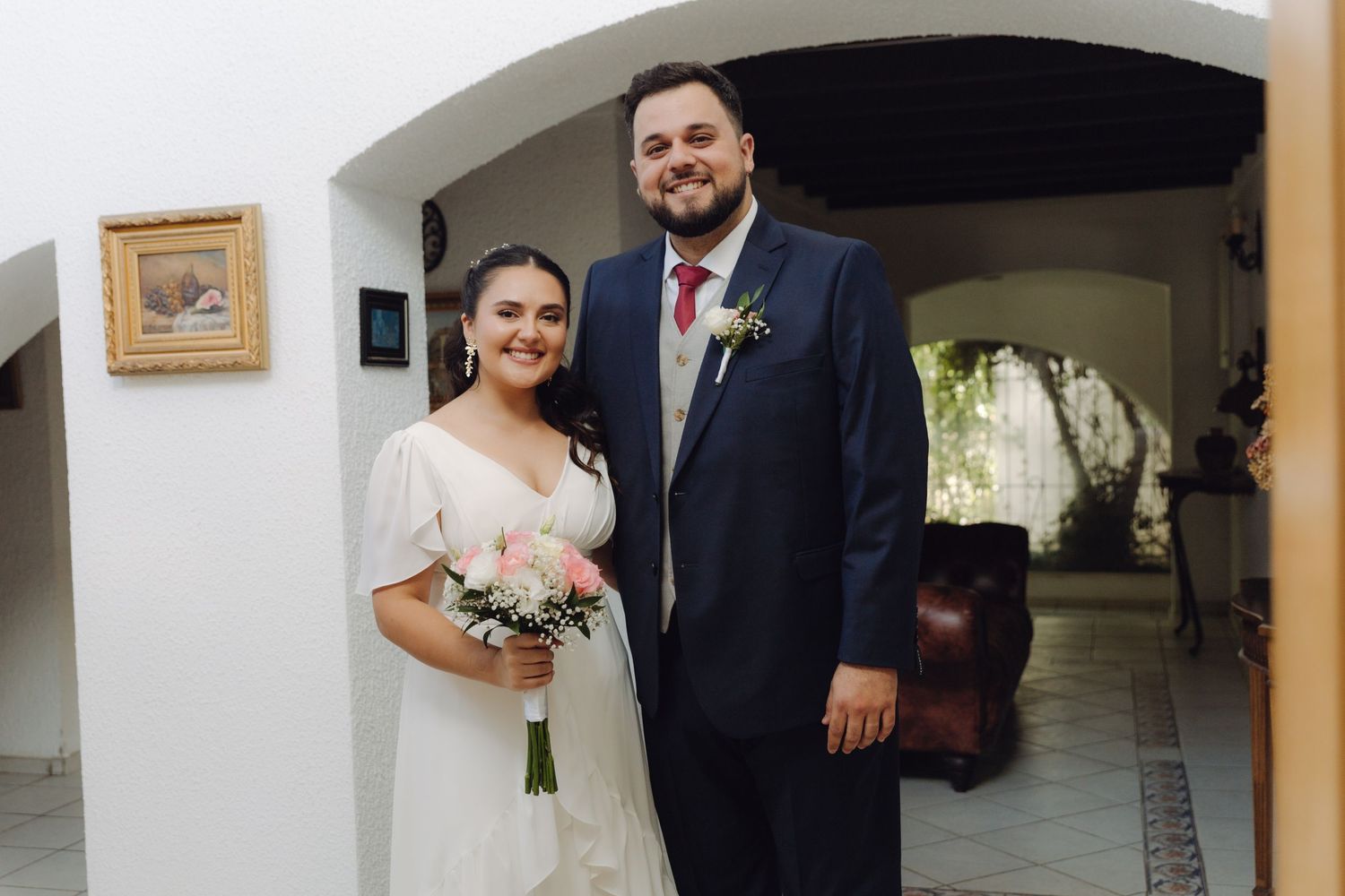 Smiling bride and groom pose together indoors, bride holding pink bouquet in white wedding dress.