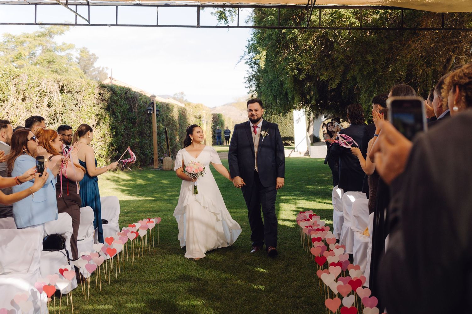Bride and groom walk down outdoor aisle lined with pink flowers as guests applaud.
