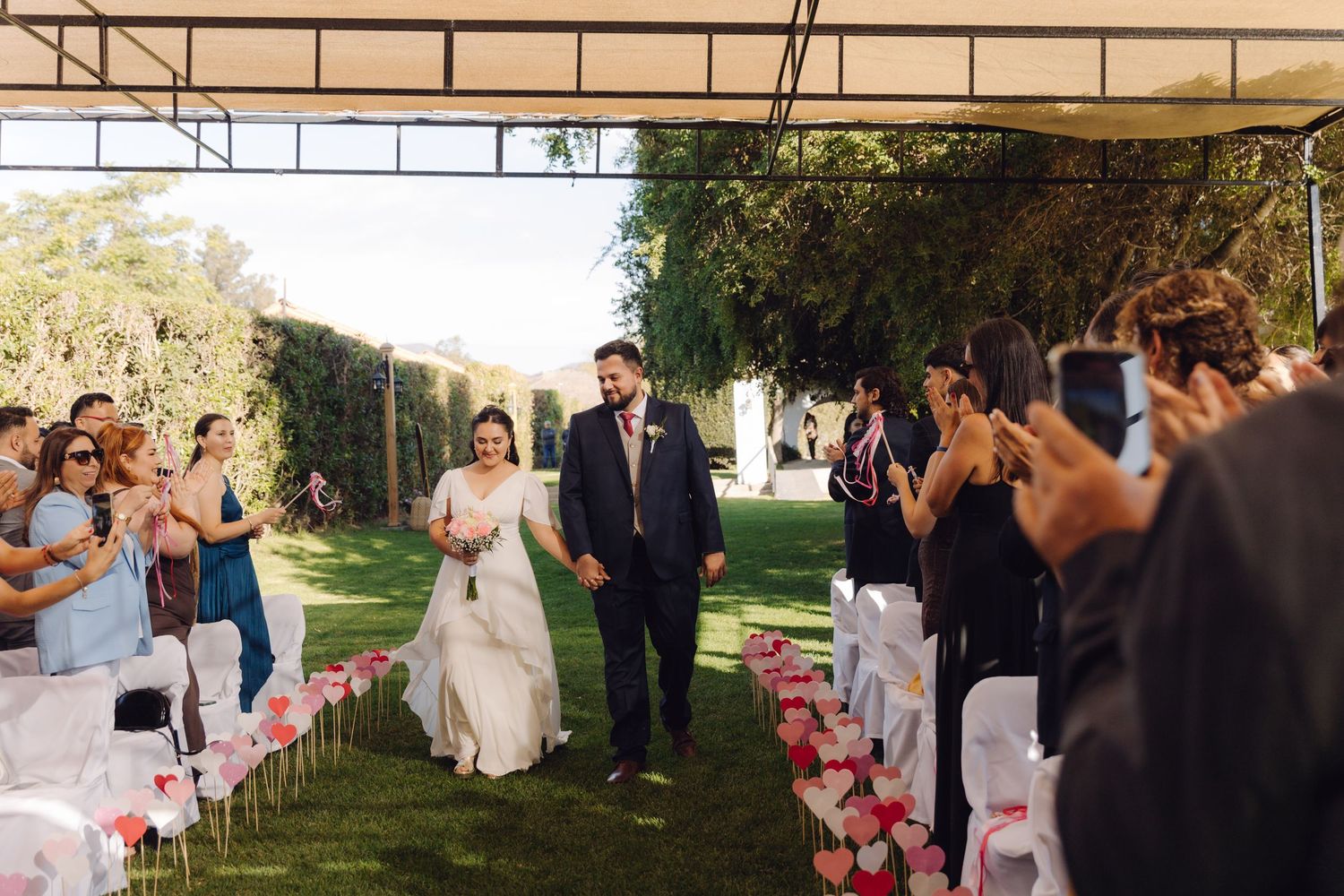 Newlyweds walk hand in hand down flower-lined outdoor aisle as guests photograph the moment.