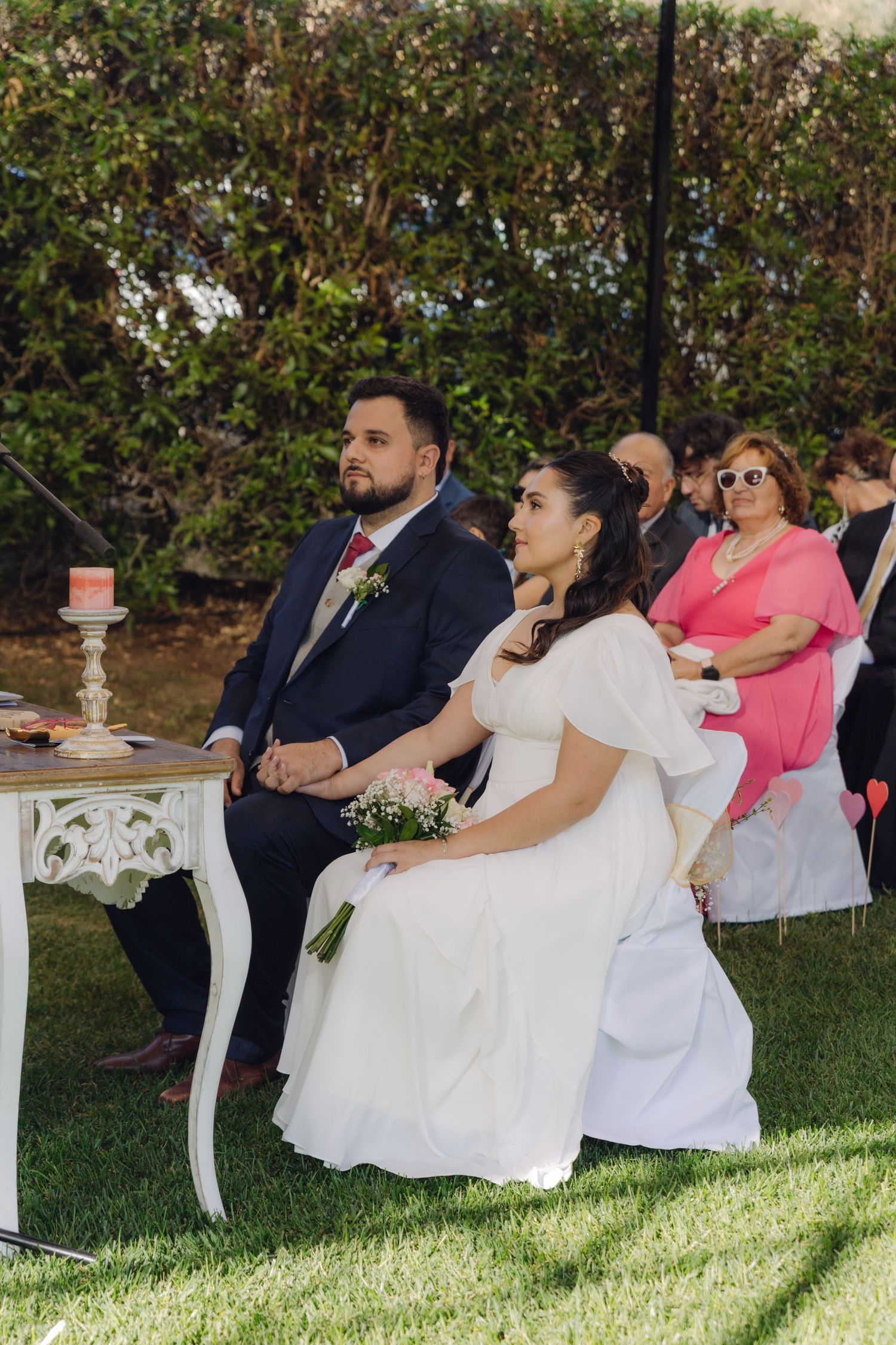 Bride in white dress and groom in navy suit seated at ornate table during outdoor wedding ceremony.