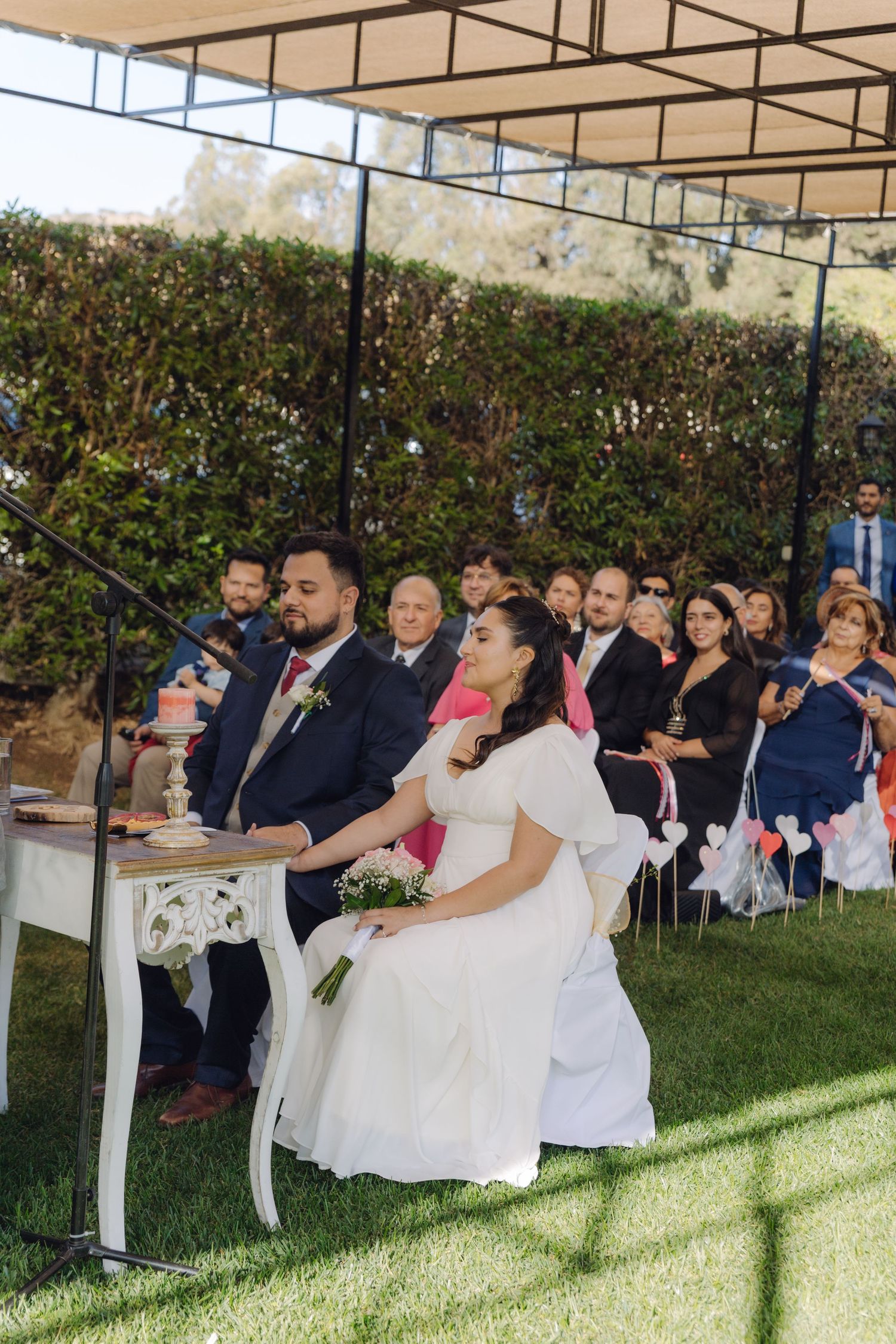 Bride and groom seated at decorated table during outdoor wedding ceremony with guests in background.