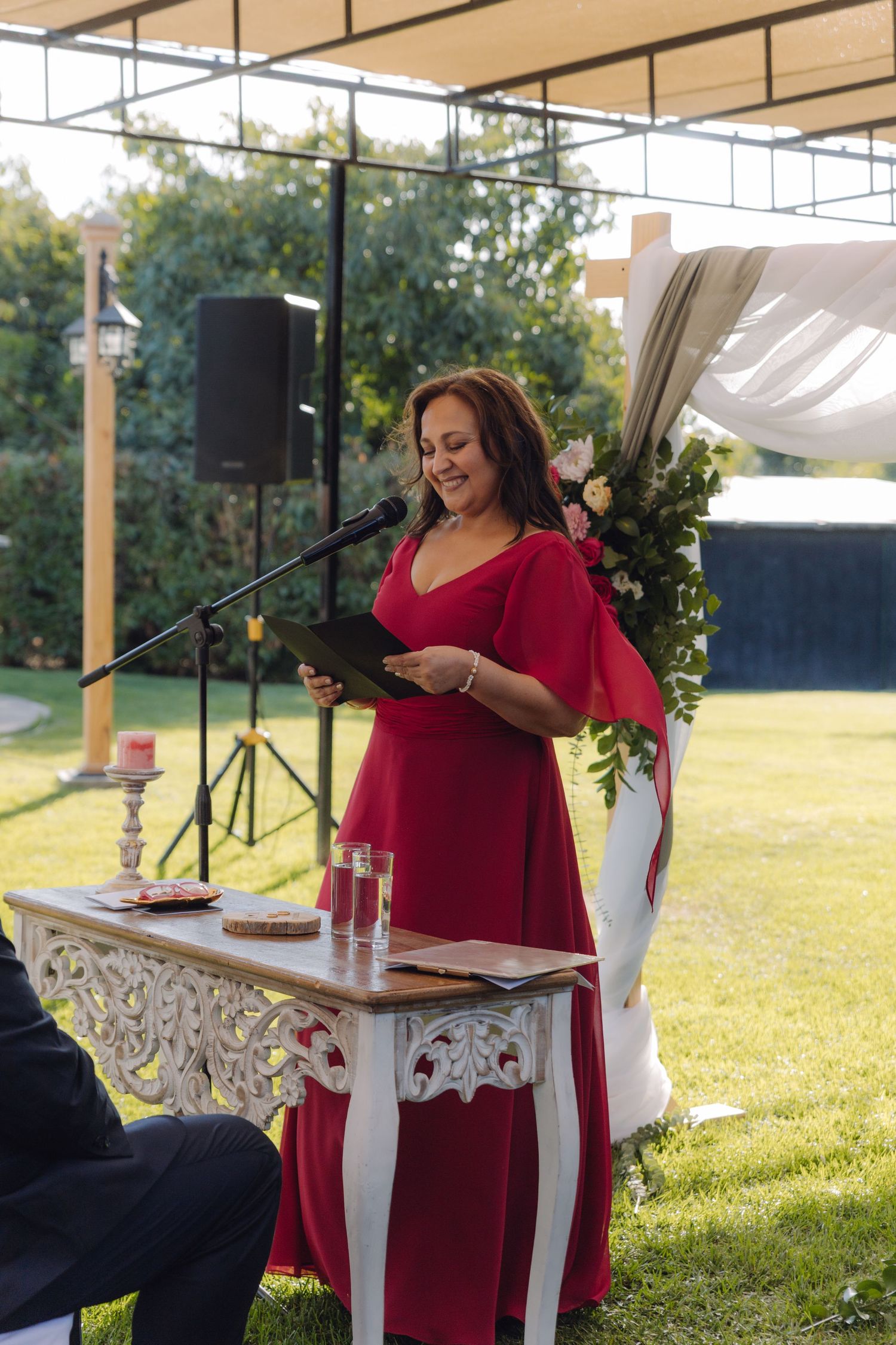 Woman in red dress reads from tablet at microphone during outdoor wedding ceremony at decorated altar.