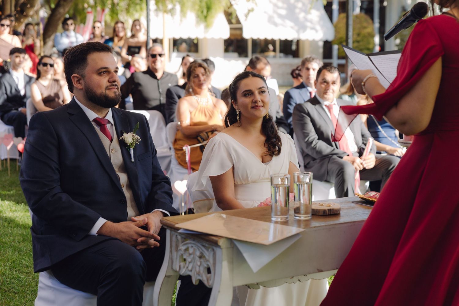 Smiling bride and groom listen to officiant in red dress during outdoor garden wedding ceremony.