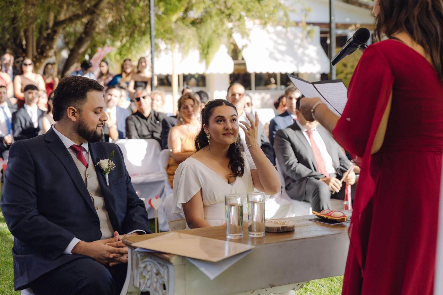Emotional bride wipes tear while groom watches officiant in red dress at outdoor wedding ceremony.