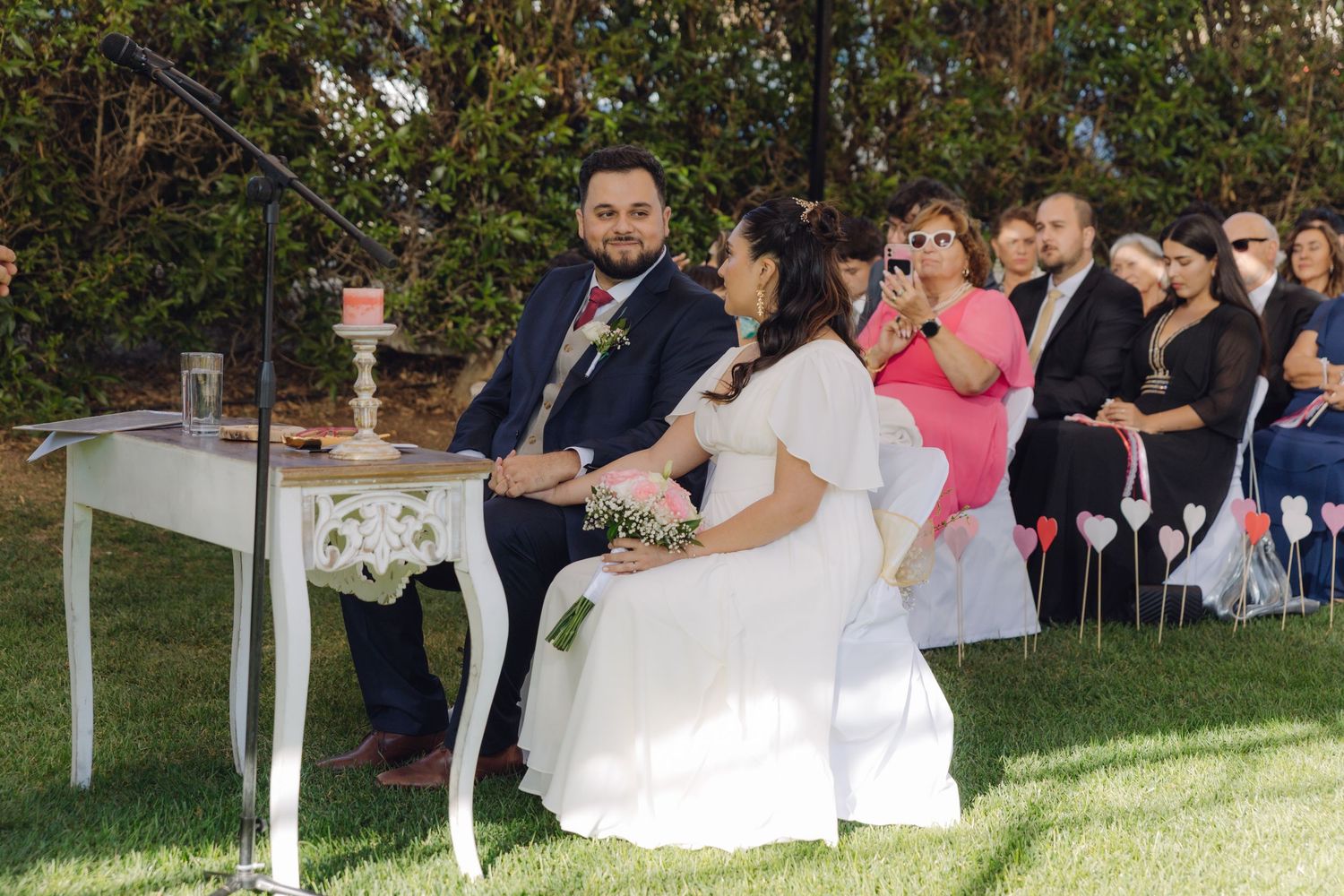 Bride holding pink bouquet and groom in navy suit seated together at outdoor wedding ceremony.