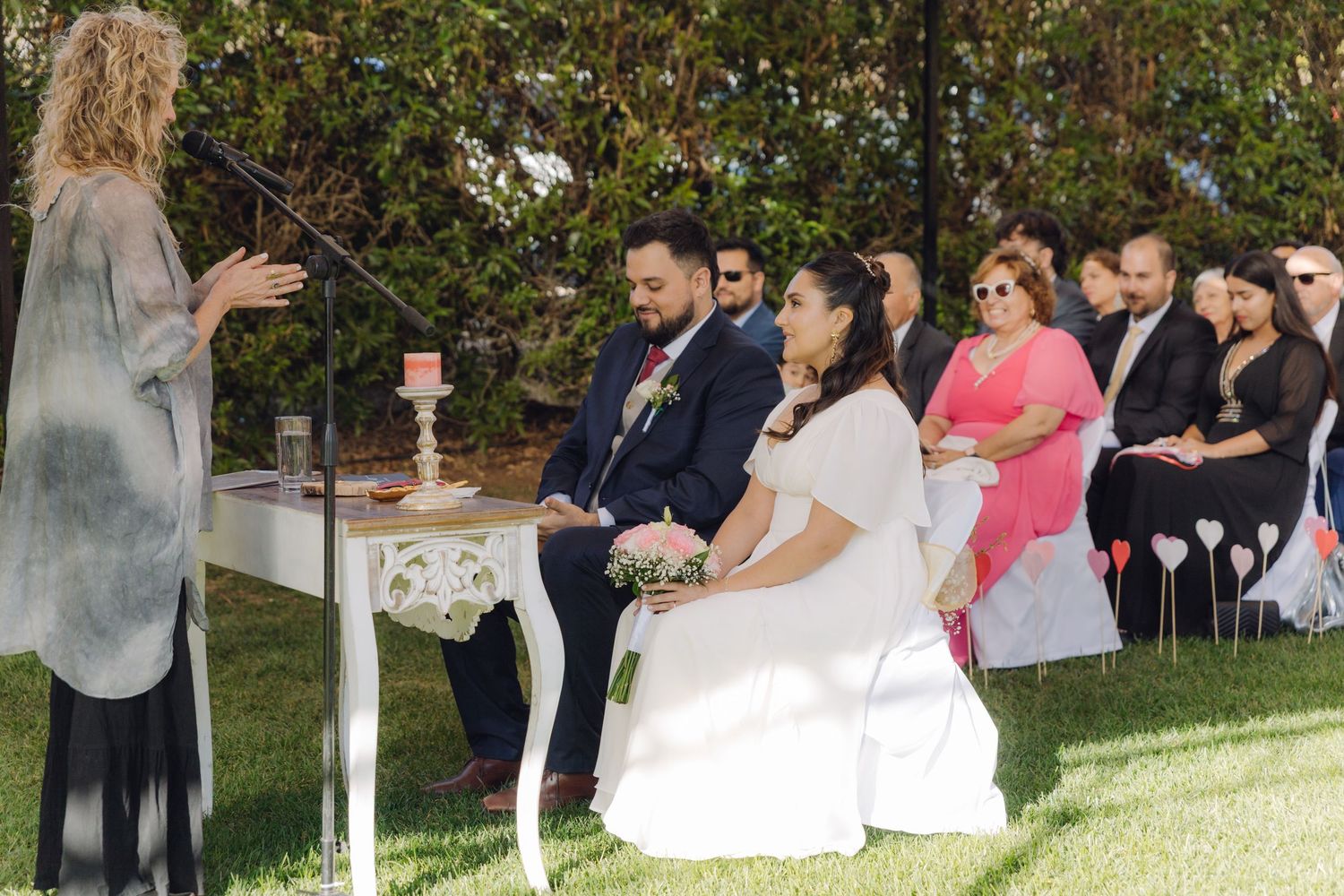 Officiant addresses bride and groom seated at ornate white table during outdoor garden wedding.