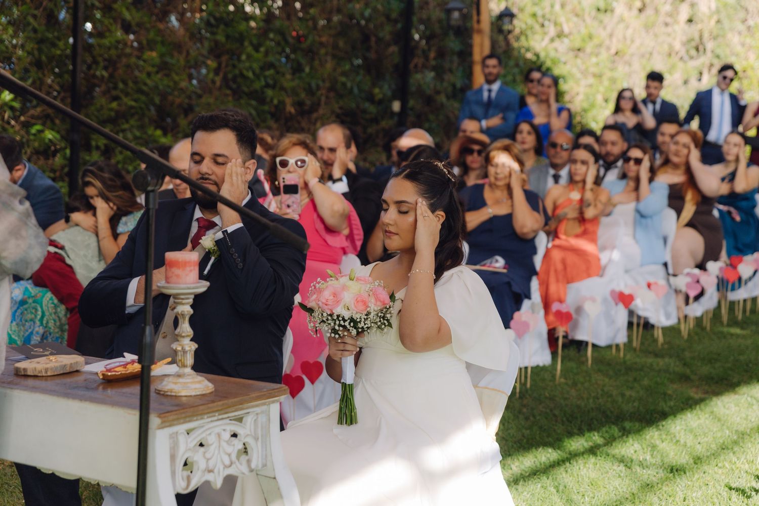 Bride covers ears playfully while groom speaks into microphone at outdoor wedding ceremony.