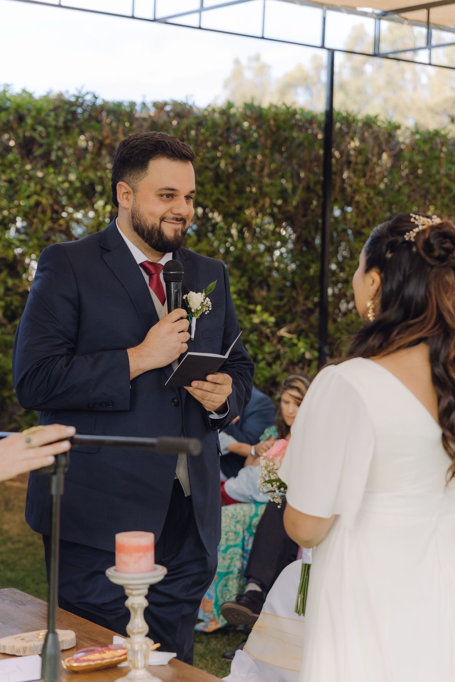 Groom in navy suit reads wedding vows aloud at outdoor ceremony while bride in white dress listens.