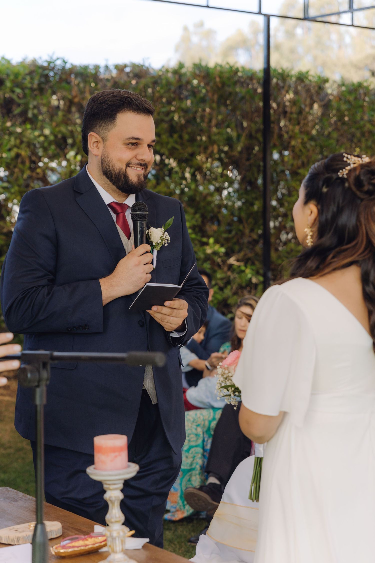 Smiling groom holds microphone and vow booklet during outdoor wedding ceremony with bride facing him.