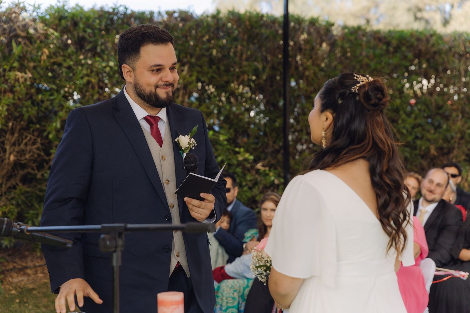 Groom and bride exchange smiles during outdoor garden wedding ceremony with guests seated behind them.