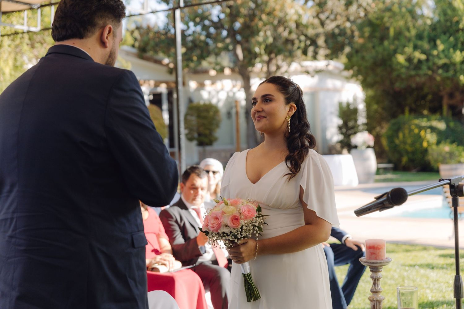 Radiant bride holds pink bouquet and smiles at groom during sunny outdoor wedding ceremony.