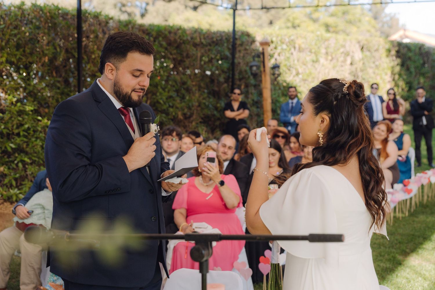 Groom speaks vows into microphone as emotional bride wipes tears at outdoor wedding ceremony.