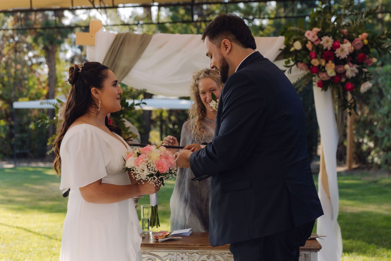 Officiant presides as groom places ring on brides hand during outdoor garden wedding ceremony.