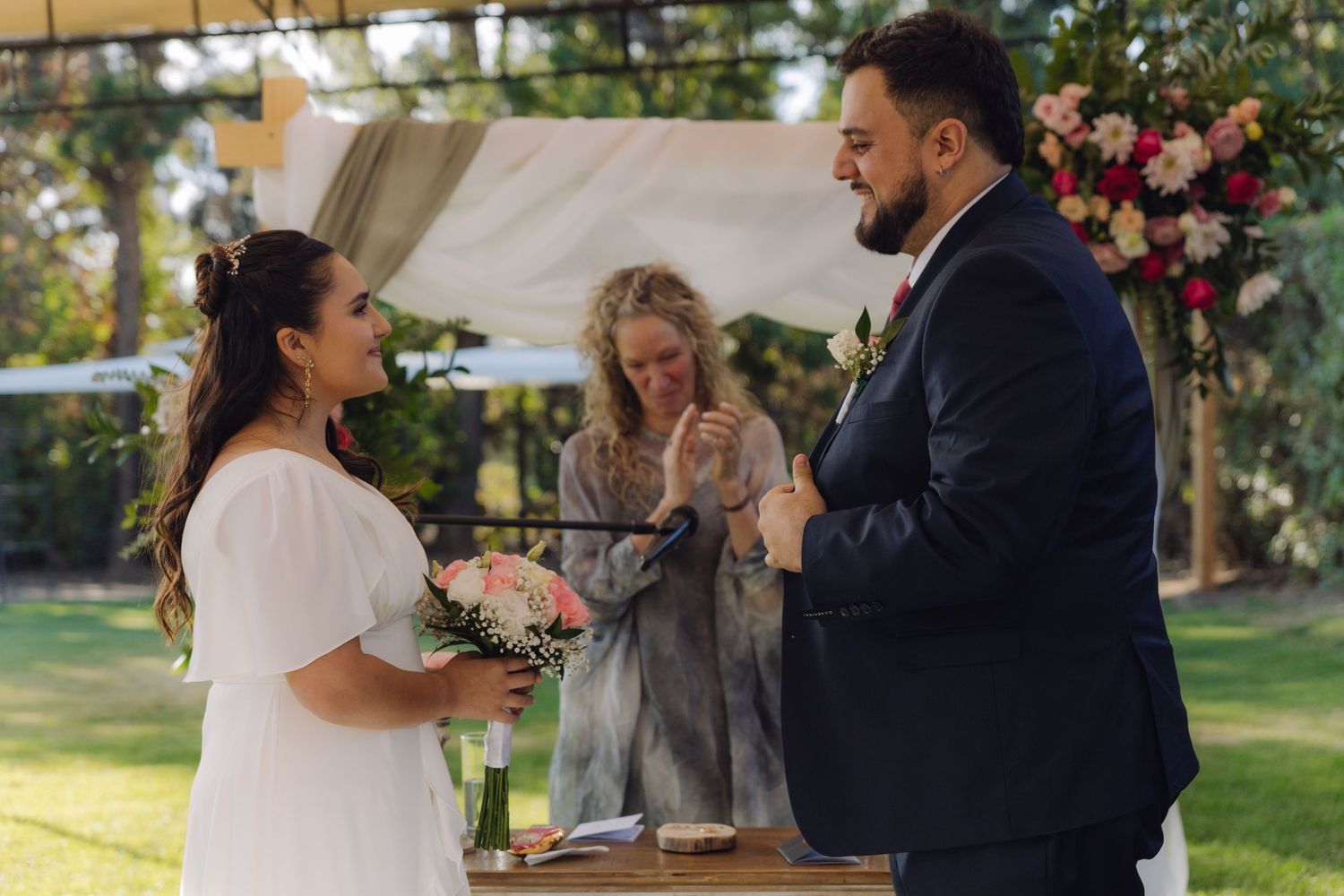 Bride and groom stand at altar with blonde officiant during elegant outdoor garden wedding ceremony.