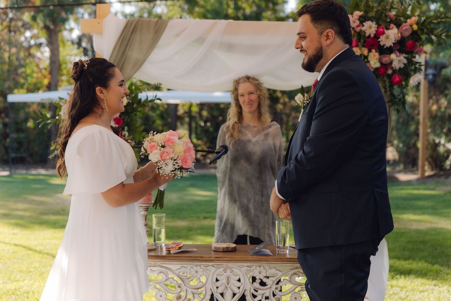 Bride laughs joyfully holding pink bouquet as groom smiles during outdoor garden wedding ceremony.