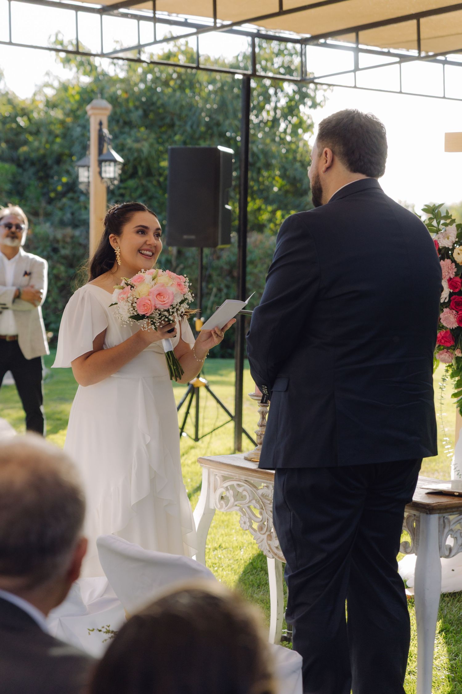 Beaming bride holds pink bouquet while groom reads vows at sunlit outdoor wedding ceremony.