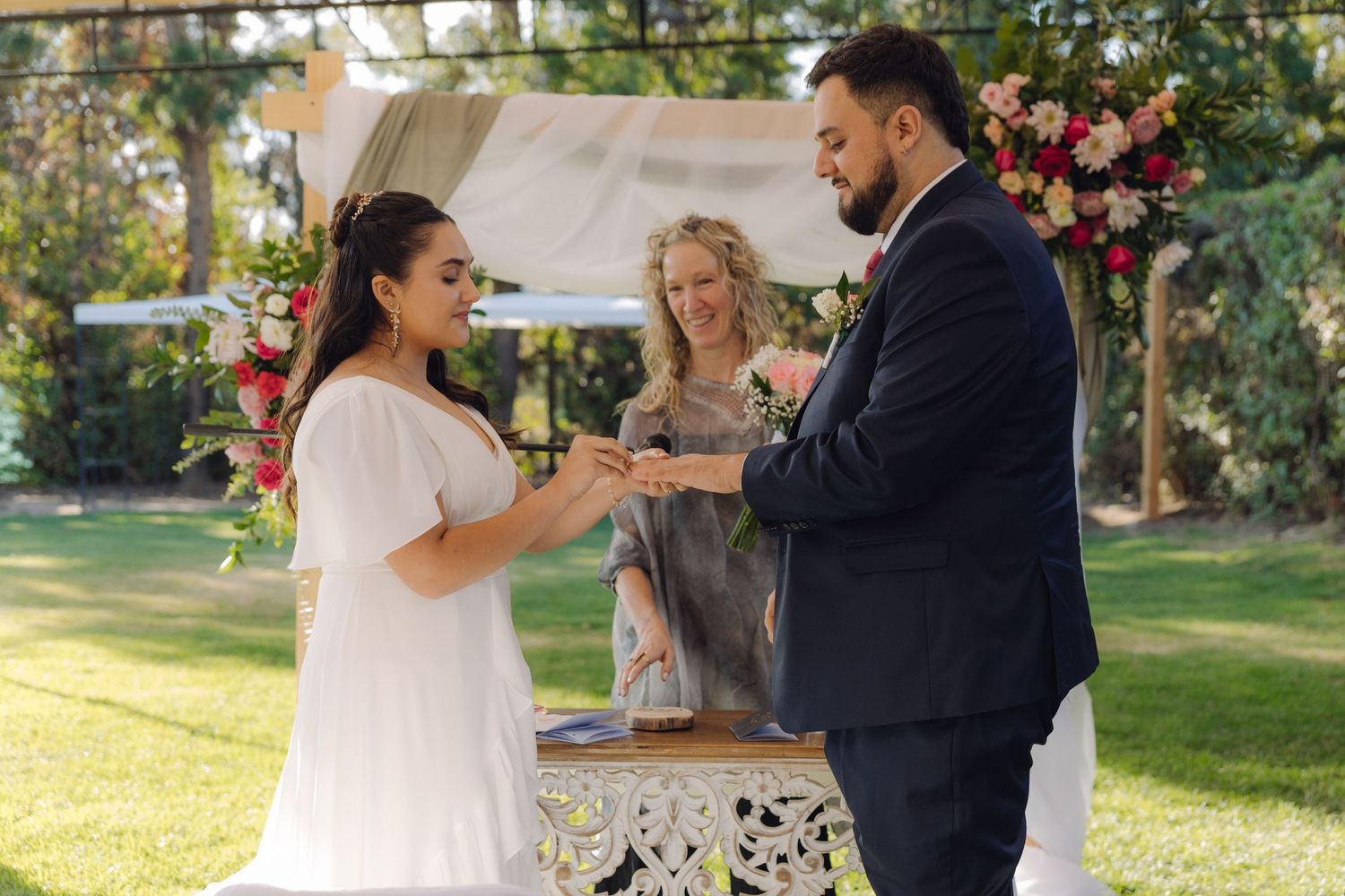 Groom places wedding ring on brides finger as smiling officiant witnesses outdoor garden ceremony.