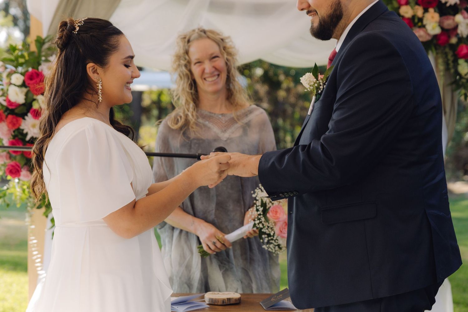 Bride smiles as officiant oversees ring exchange with groom at floral outdoor wedding ceremony.